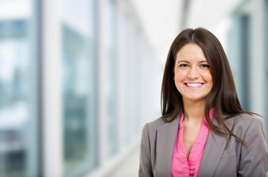 Modern ofis koridorundaki kendine güvenen iş kadını profesyonellik ve başarı yayıyor, zarif bir takım elbise içinde mutluluk ve iyimserlik yayıyor, pencereden dışarı bakıyor.