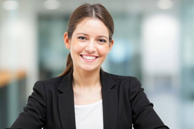 Genç bir iş kadını modern bir ofis binasında otururken gülümsüyor. O siyah bir ceket ve beyaz bir gömlek giyiyor.