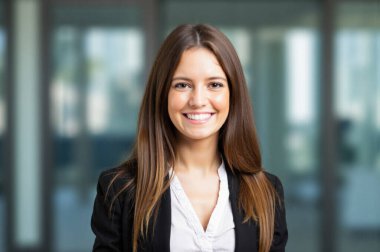 Modern ofis ortamında kendine güvenen genç bir iş kadını şık siyah ceket ve beyaz gömleğiyle ışıl ışıl gülümsüyor başarı ve kararlılık saçıyor. İş projeleri için mükemmel