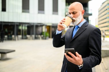 Üst düzey kel yönetici, beyaz sakallı, dışarıda kahve içerken akıllı telefonunu kullanıyor.