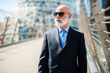 Sakallı, kendinden emin kıdemli işadamı güneşli bir günde şehirde yürüyor. Takım elbise ve kravat takıyor ve yan tarafa bakıyor.