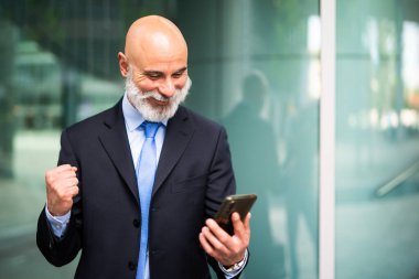 Olgun iş adamı ofis binasının önünde dikiliyor, akıllı telefonuna bakıyor ve gülümsüyor. Zafer jesti olarak yumruğunu kaldırıyor.