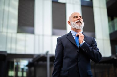 Kıdemli işadamı kravatını düzeltirken modern bir ofis binasının önünde duruyor, güven ve deneyim duygusu aktarıyor.