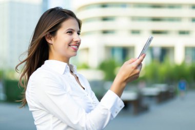 Şehirdeki cep telefonunu kullanarak gülümseyen genç iş kadını, güneşli bir günün keyfini çıkarırken aynı zamanda çevrimiçi olarak başkalarıyla iletişim kuruyor ve şehir ortamında modern teknolojiyi kucaklıyor.