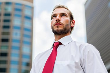 Gıcır gıcır beyaz bir gömlek ve canlı kırmızı bir kravat takan genç bir yönetici, gökyüzüne bakıyor, modern ofis binalarıyla çevrili, hareketli bir şehir manzarası içinde
