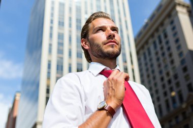 Genç işadamı, güneşli bir günde şehirde gökdelenlerin önünde dikilirken kırmızı kravatını düzeltiyor. Kendinden emin ve başarılı görünüyor.