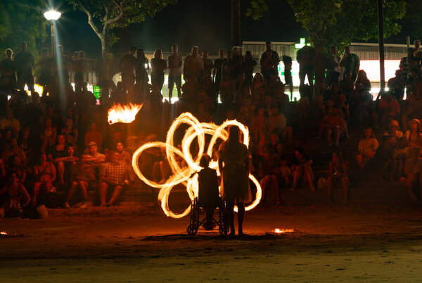 A picture of the fire show at the Ao Nang Beach, at night. Here, the performer is in a wheelchair, and plays with the fire around a woman from the crowd.