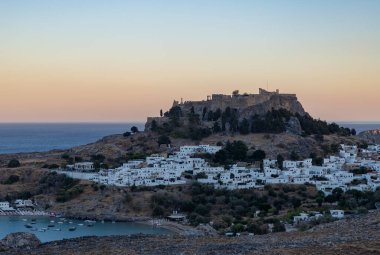 Lindos kasabasının ve gün batımında çevreleyen manzaranın resmi..
