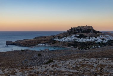 Lindos kasabasının ve gün batımında çevreleyen manzaranın resmi..