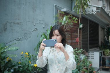 Akıllı telefon tutan genç bir kadın, açık hava bahçesi, günlük kıyafetler, doğal ışık, odaklanmış.