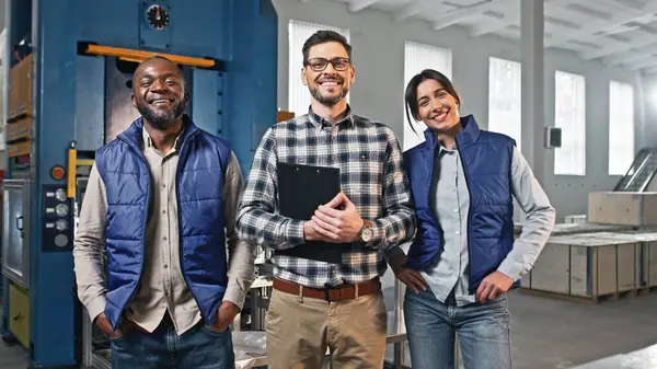 カメラを直接見ながら笑顔で笑っている3人の多民族の姿 製造工場の2人のエンジニアの間に立っているクリップボードを持つハンサムマネージャー 彼らの仕事を楽しむ ロイヤリティフリーのストック写真