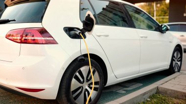 Kamera şarj olurken dışarıda park etmiş elektrikli bir arabadan uzaklaşır. EV gelecek teknolojisi. Sıfır emisyon otomatik, çevre dostu araç. Caddedeki şarj istasyonunda beyaz elektroaraba. Eklenti komutu
