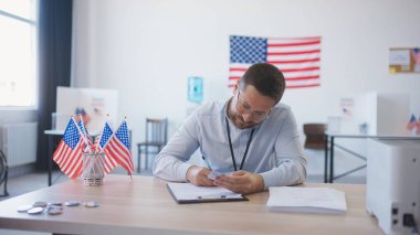 Masasında gözlüklü, zeki beyaz bir erkek oturuyor. Kamera arkasındaki birinden kimlik kabul etmek ve karttan bilgi almak. Masada minyatür Amerikan bayrakları var. Halk cumhurbaşkanına oy veriyor.