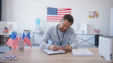 Masasında gözlüklü, zeki beyaz bir erkek oturuyor. Kamera arkasındaki birinden kimlik kabul etmek ve karttan bilgi almak. Masada minyatür Amerikan bayrakları var. Halk cumhurbaşkanına oy veriyor.