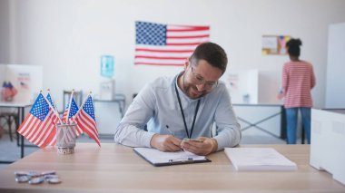 Masasında gözlüklü beyaz bir erkek oturuyor ve kadından kimlik kabul ediyor. Bilgileri kontrol ediyor ve kadınlara oy pusulası ve yuvarlak rozet veriyor. Masada Amerikan bayrakları var. Cumhurbaşkanlığı seçimleri.