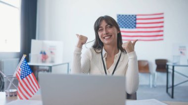 Ofiste oturan mutlu bir kız ya da Amerikan bayraklı bir seçim merkezi. Yaygın bir şekilde gülümsüyor ve kameraya başparmaklarını gösteriyor. Başkanlık seçimlerinin tadını çıkarıyorum. İnsanları oy kullanma yerine gitmeye teşvik etmek.