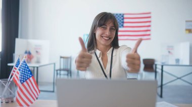 Ofiste oturan mutlu bir kız ya da Amerikan bayraklı bir seçim merkezi. Yaygın bir şekilde gülümsüyor ve kameraya başparmaklarını gösteriyor. Başkanlık seçimlerinin tadını çıkarıyorum. İnsanları oy kullanma yerine gitmeye teşvik etmek.