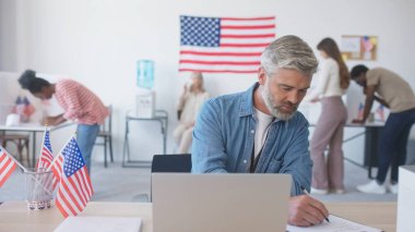 Dizüstü bilgisayar kullanan çalışkan bir adam. Belgelerle çalışıyorum. Seçimlerde oy verirken önemli verilerin karşılaştırılması. Amerikan vatandaşları arka planda cumhurbaşkanı adayı hakkında karar veriyorlar.