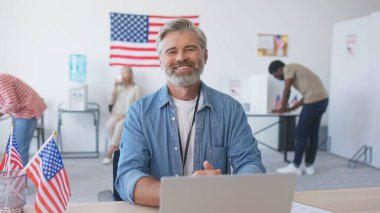 Dizüstü bilgisayar kullanan çalışkan bir adam. Belgelerle çalışıyorum. Seçimlerde oy verirken önemli verilerin karşılaştırılması. Amerikan vatandaşları arka planda cumhurbaşkanı adayı hakkında karar veriyorlar.