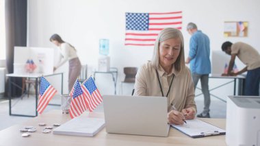 Amerikan vatandaşları yerel oy kullanma merkezini ziyaret ediyor. Dizüstü bilgisayarla masada otururken seçmenlerle buluşan gülümseyen kadın. Anket görevlisi belgeleri kontrol ediyor ve oy verme formu veriyor. Başkan seçiliyor.