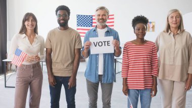 Yan yana duran çok ırklı, günlük kıyafetler giymiş insanlar. Amerikan ofisi ya da eyalet oy kullanma merkezi. Elinde 