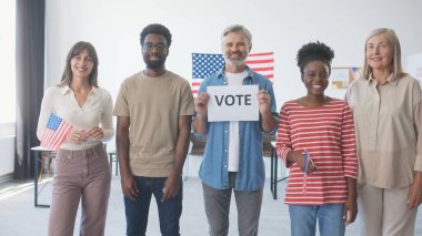 Yan yana duran çok ırklı, günlük kıyafetler giymiş insanlar. Amerikan ofisi ya da eyalet oy kullanma merkezi. Elinde 