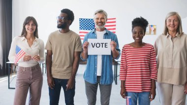 Bir grup insan sandık merkezinde sırada bekliyor. Duvarda asılı duran Amerika Birleşik Devletleri bayrağında. İnsanlar gülümsüyor. Seçimlerini oy pusulasından yaptıkları için mutlular. Vatanseverler ülkesi.