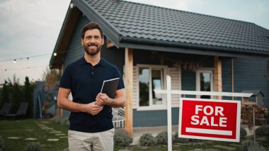 Beyaz metin ile kırmızı tabelasının yanında duran yakışıklı beyaz erkek satılık. Sakallı adam kameraya bakarken gülümsüyor. Not defterini tutuyorum. Arka planda bulanık küçük bir bina. Kırsal.