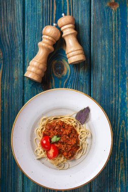 Beyaz tabakta spagetti bolonez makarna, tuz, biber, peynir ve domatesler mavi ahşap arka planda.