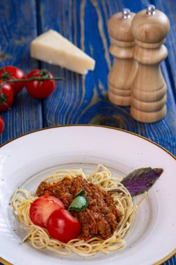 Beyaz tabakta spagetti bolonez makarna, tuz, biber, peynir ve domates mavi ahşap arka planda.