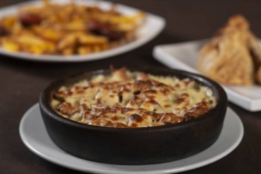 Erimiş peynirle kaplanmış mantar ve et güveci masada siyah bir tabakta servis edilir.