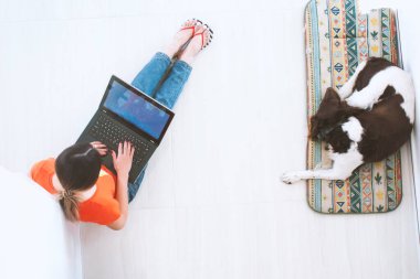 Genç bir kadın köpeğiyle yerde yatıyor ve bir dizüstü bilgisayar üzerinde çalışıyor.