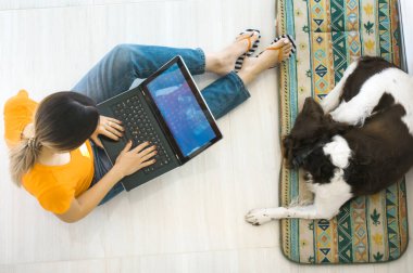 Genç bir kadın köpeğiyle yerde yatıyor ve bir dizüstü bilgisayar üzerinde çalışıyor.