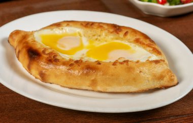 Adjar Khachapuri. Tahta arka planda beyaz tabakta yumurta ve peynirli Gürcistan turtası..