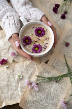 Bir fincan kapuçino tutan kadın eller ve arka planda çiçekler.