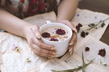 Bir fincan kapuçino tutan kadın eller ve arka planda çiçekler.