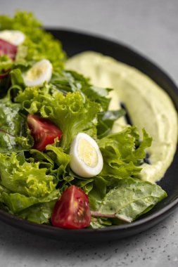 Fresh green salad with quail eggs, cherry tomatoes and guacamole in black plate on grey background