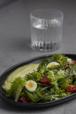 Fresh green salad with quail eggs, cherry tomatoes and guacamole in black plate on grey background