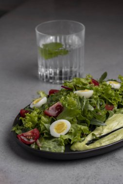Fresh green salad with quail eggs, cherry tomatoes and guacamole in black plate on grey background