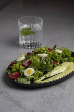 Fresh green salad with quail eggs, cherry tomatoes and guacamole in black plate on grey background