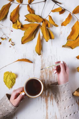 Sarı yapraklı, sonbahar kompozisyonu olan, pejmürde masa üzerinde çay ve termometre tutan kadınların üst görüntüsü.