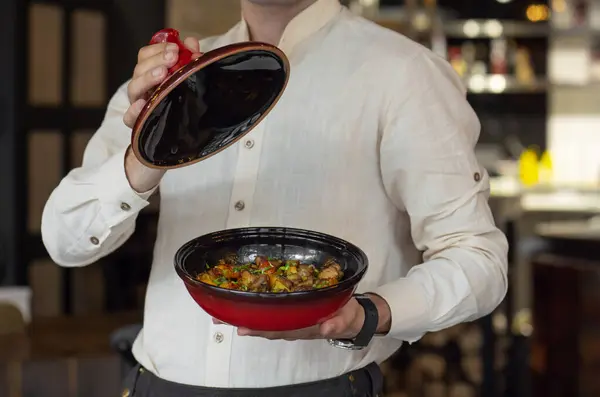Waiter serving a dish with fried meat with vegetables in a ceramic bowl on a light background