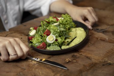 Person eating Fresh green salad with quail eggs, cherry tomatoes and guacamole in black plate on grey background