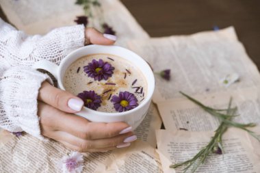 Bir fincan kapuçino tutan kadın eller ve arka planda çiçekler.