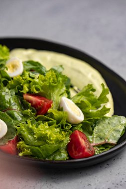 Fresh green salad with quail eggs, cherry tomatoes and guacamole in black plate on grey background