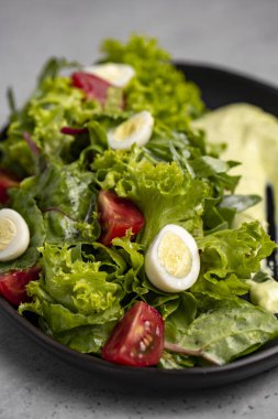Fresh green salad with quail eggs, cherry tomatoes and guacamole in black plate on grey background