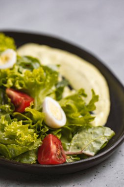 Fresh green salad with quail eggs, cherry tomatoes and guacamole in black plate on grey background