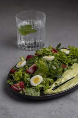 Fresh green salad with quail eggs, cherry tomatoes and guacamole in black plate on grey background