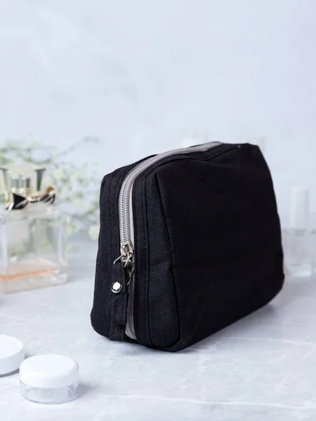 stock image Black cosmetic bag  on a stone table, with flowers and perfume in the background