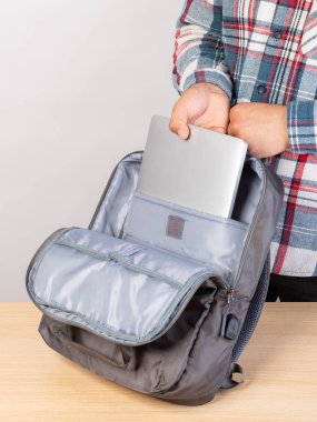 Ekose gömlekli bir adam elini gri bir dizüstü bilgisayara koyup şık gri bir sırt çantası takıyor..